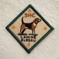A diamond-shaped embroidered patch with a green border. It features a brown and black beagle standing facing left, with the text "SHC" above and "CANINE BUREAU" below the dog. The patch has four small stars at the corners.