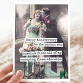 An anniversary card with a vintage photo of a man in a suit embracing a woman in a light blue dress, both holding flowers. Text on the right side of the card reads: "Happy Anniversary to the person who somehow finds all of my annoying flaws adorable." The card is resting on a textured cream-coloured fabric with some wispy dried grasses.