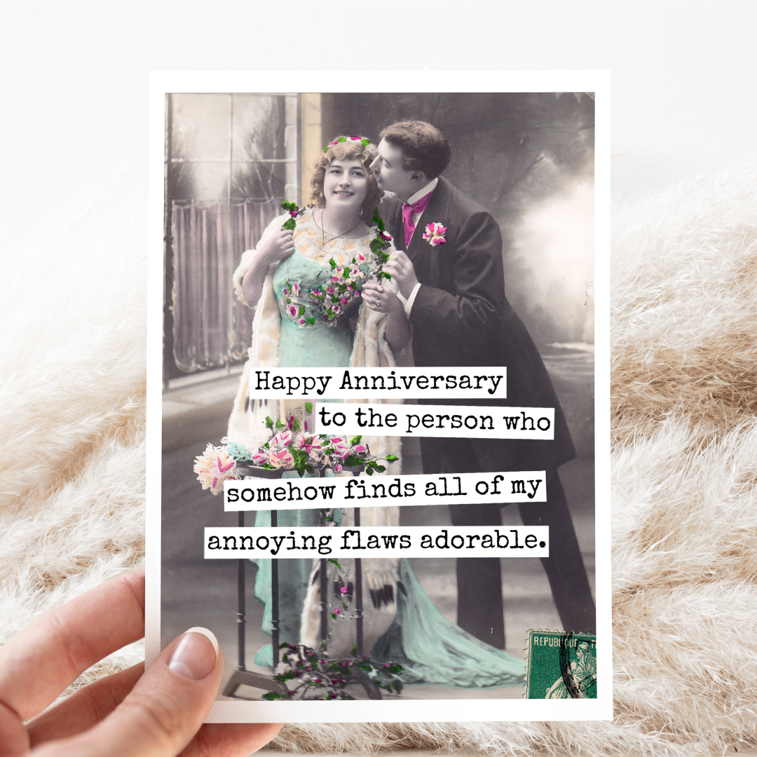 An anniversary card with a vintage photo of a man in a suit embracing a woman in a light blue dress, both holding flowers. Text on the right side of the card reads: "Happy Anniversary to the person who somehow finds all of my annoying flaws adorable." The card is resting on a textured cream-coloured fabric with some wispy dried grasses.