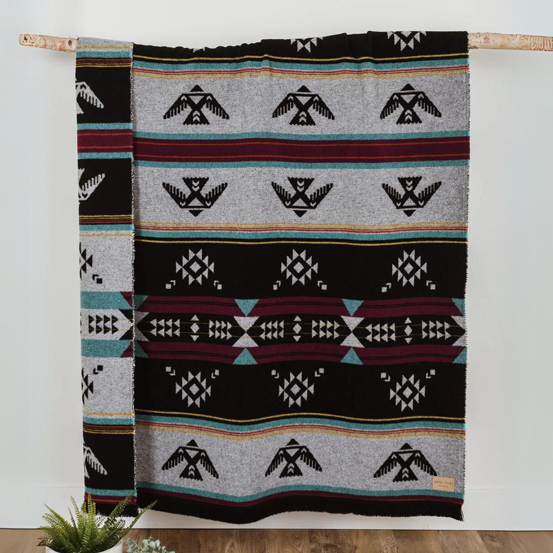 The Éclair Eco-Friendly Everyday Blanket by Mini Tipi, featuring bold Indigenous-inspired geometric patterns in black, grey, burgundy, turquoise, and yellow, hanging from a natural branch. This sustainable and stylish blanket is available in Ottawa, Ontario, Canada.