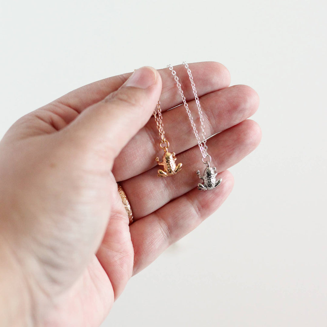 Two small frog-shaped pendants on delicate chains held in a hand against a light background.