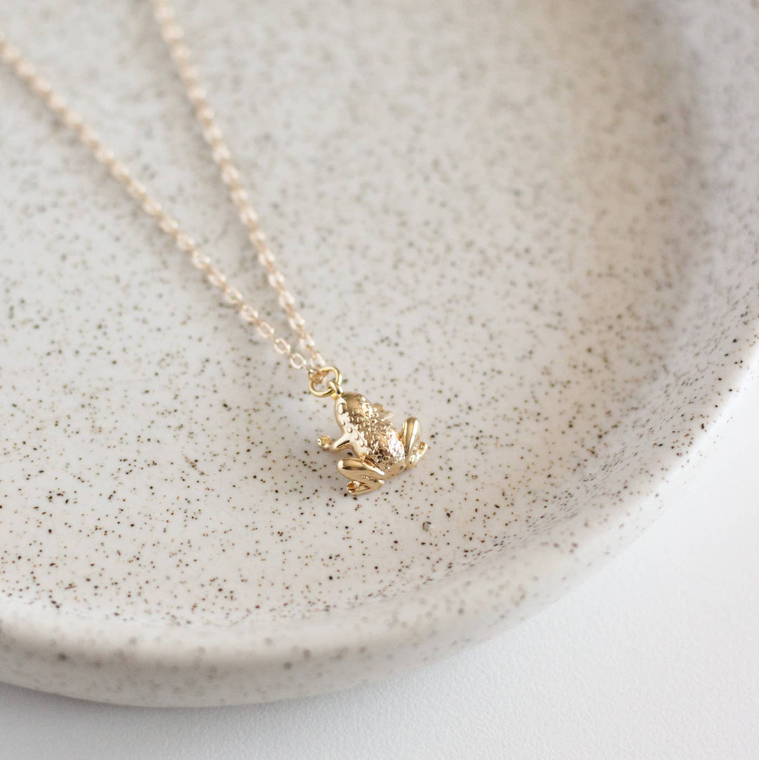 Gold frog pendant necklace on a light gray speckled ceramic plate