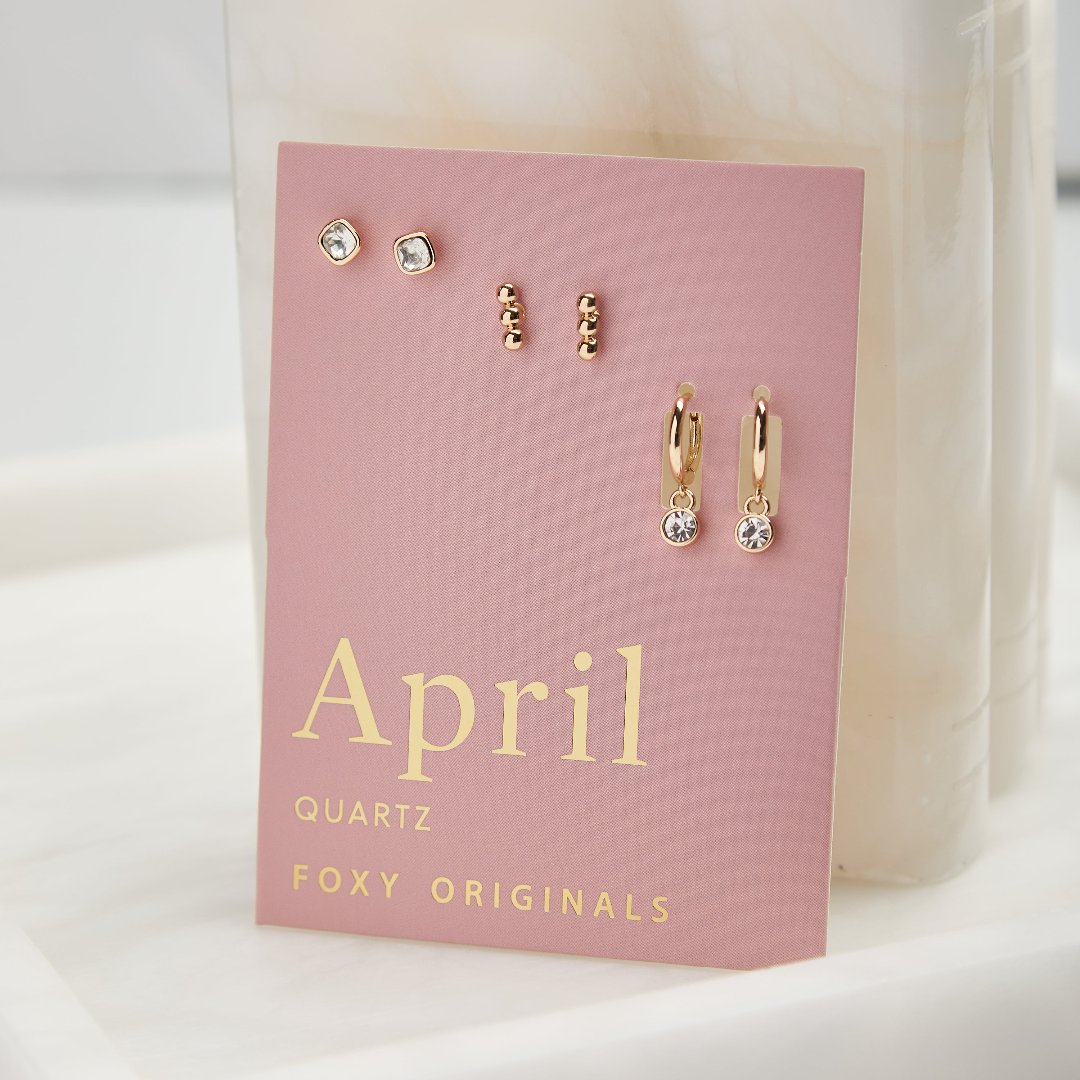 A set of three pairs of quartz earrings displayed on a pink card. The set includes a pair of stud earrings, a pair of bar earrings with small quartz beads, and a pair of hoop earrings with dangling quartz stones. The card has the month "April" and the brand name "Foxy Originals."