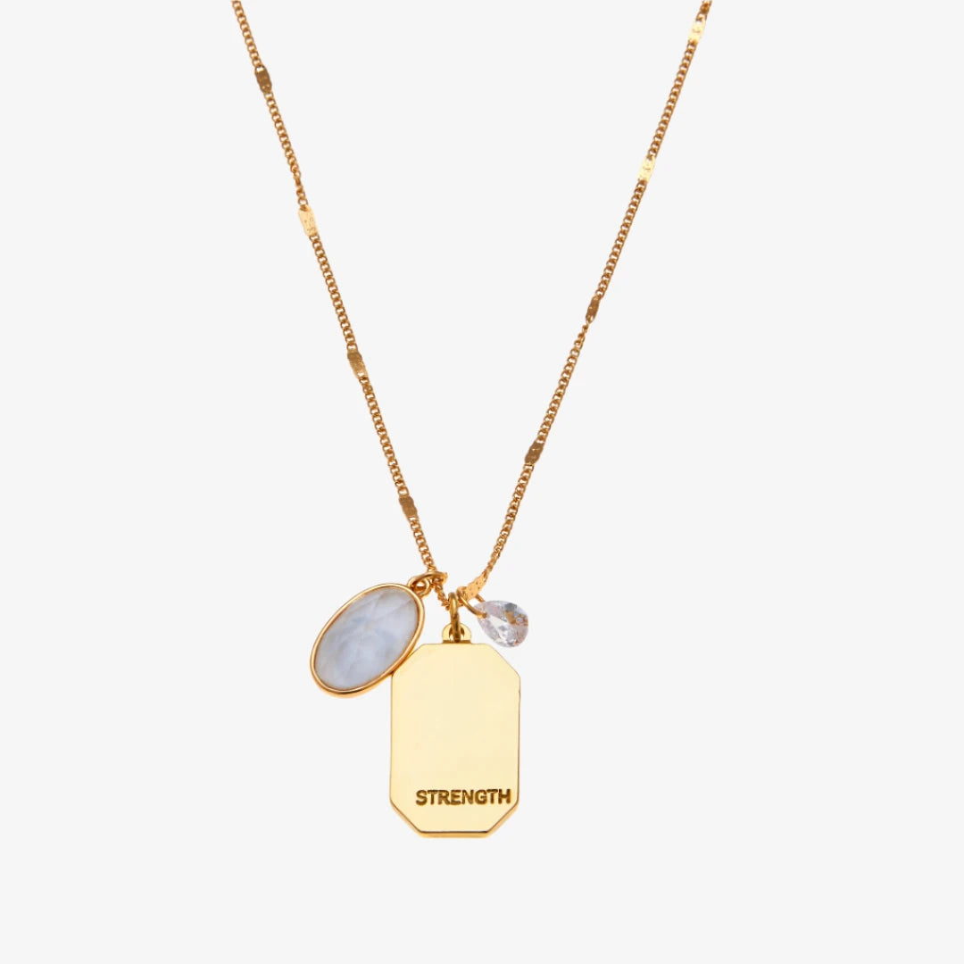 A gold necklace with a quartz stone pendant. The pendant also has the word "STRENGTH" engraved on it. The necklace is displayed on a pink card with the month "April" and the brand name "Foxy Originals."