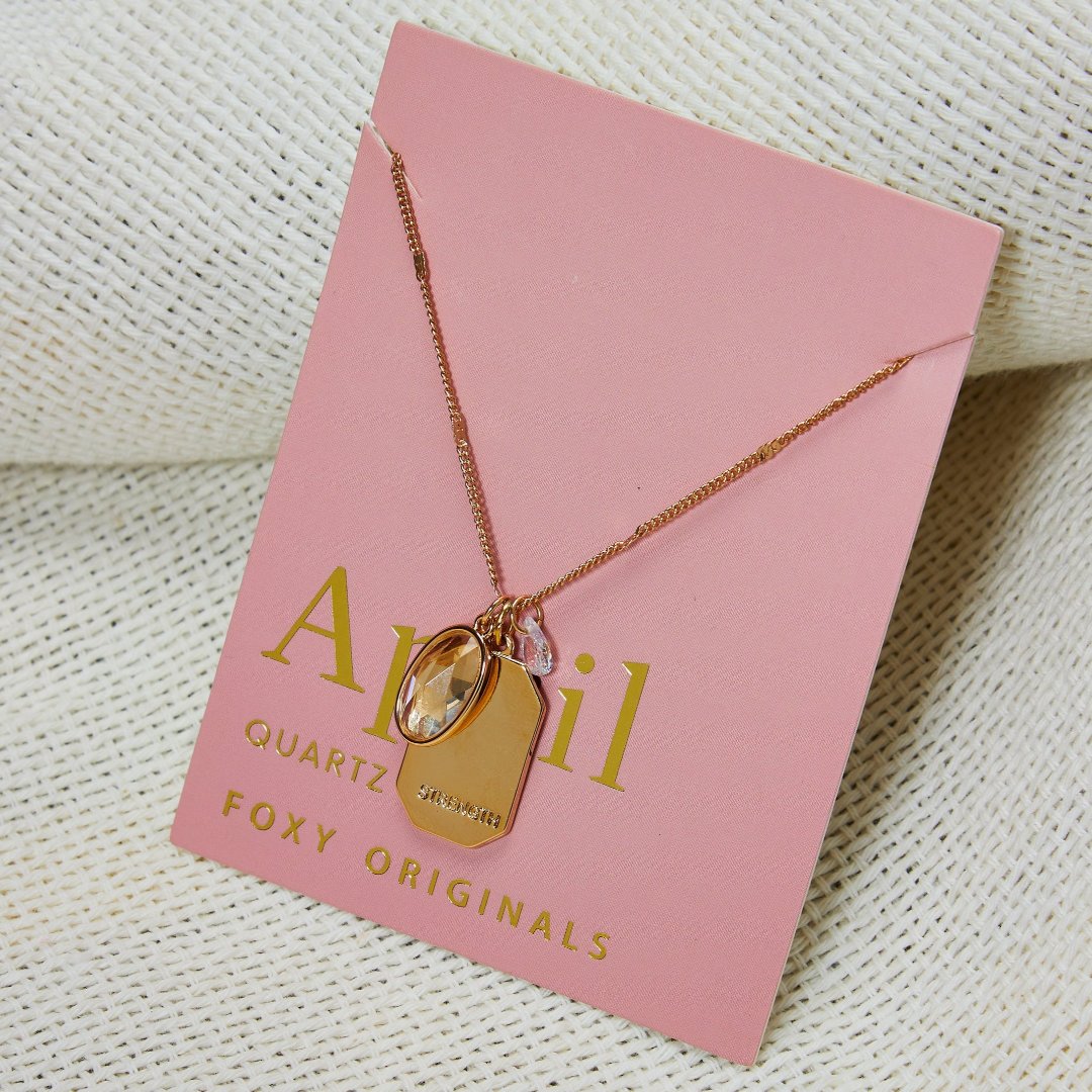 A gold necklace with a quartz stone pendant. The pendant also has the word "STRENGTH" engraved on it. The necklace is displayed on a pink card with the month "April" and the brand name "Foxy Originals."