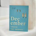 A set of three pairs of turquoise earrings displayed on a blue card. The set includes a pair of stud earrings, a pair of bar earrings with small turquoise beads, and a pair of hoop earrings with dangling turquoise stones. The card has the month "December" and the brand name "Foxy Originals."