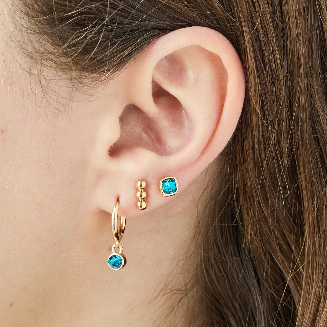 A set of three pairs of turquoise earrings displayed on a blue card. The set includes a pair of stud earrings, a pair of bar earrings with small turquoise beads, and a pair of hoop earrings with dangling turquoise stones. The card has the month "December" and the brand name "Foxy Originals."