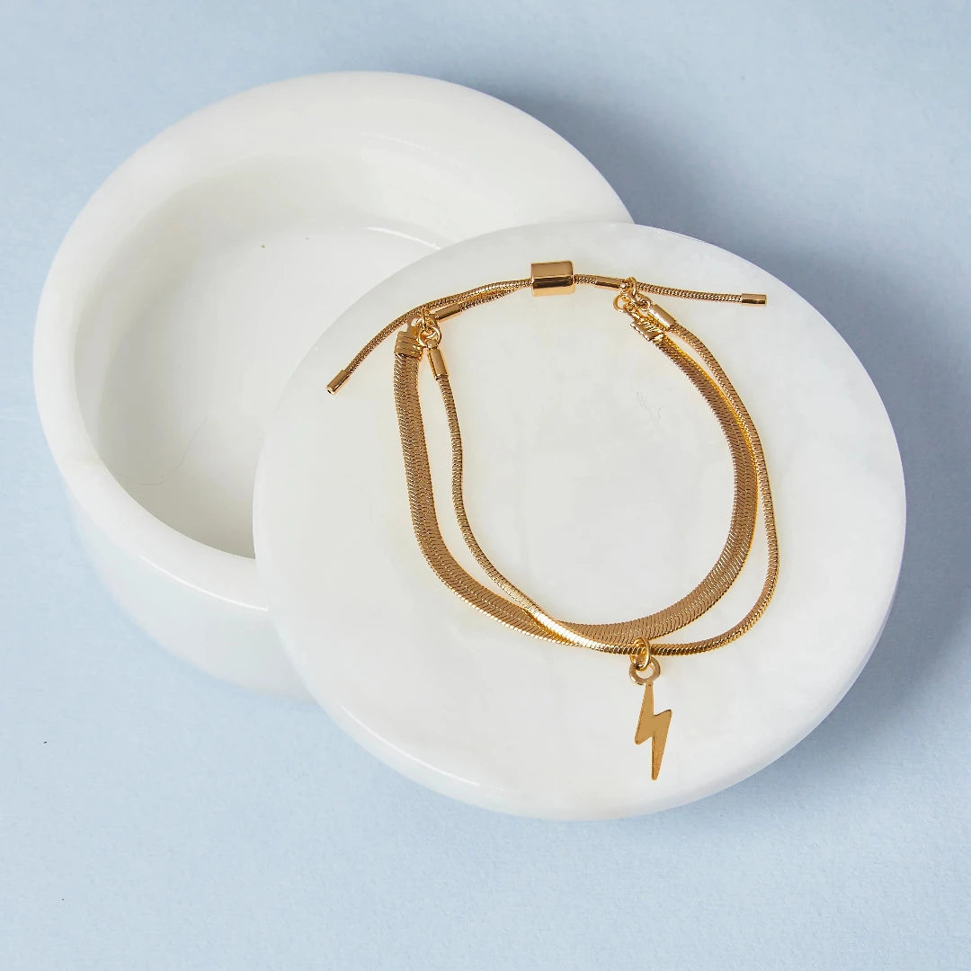 A set of two gold bracelets, one layered over the other, with a lightning bolt charm hanging from the bottom. The bracelets are displayed on a pink card with the brand name "Foxy Originals."