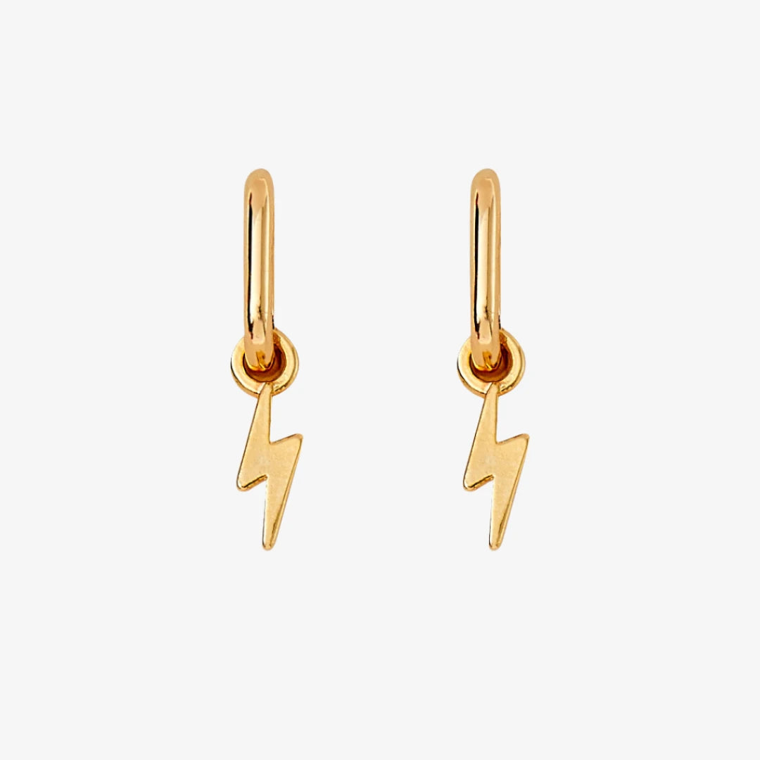 A pair of gold earrings with lightning bolt charms on a pink background. The earrings are displayed on a card with the brand name "Foxy Originals."