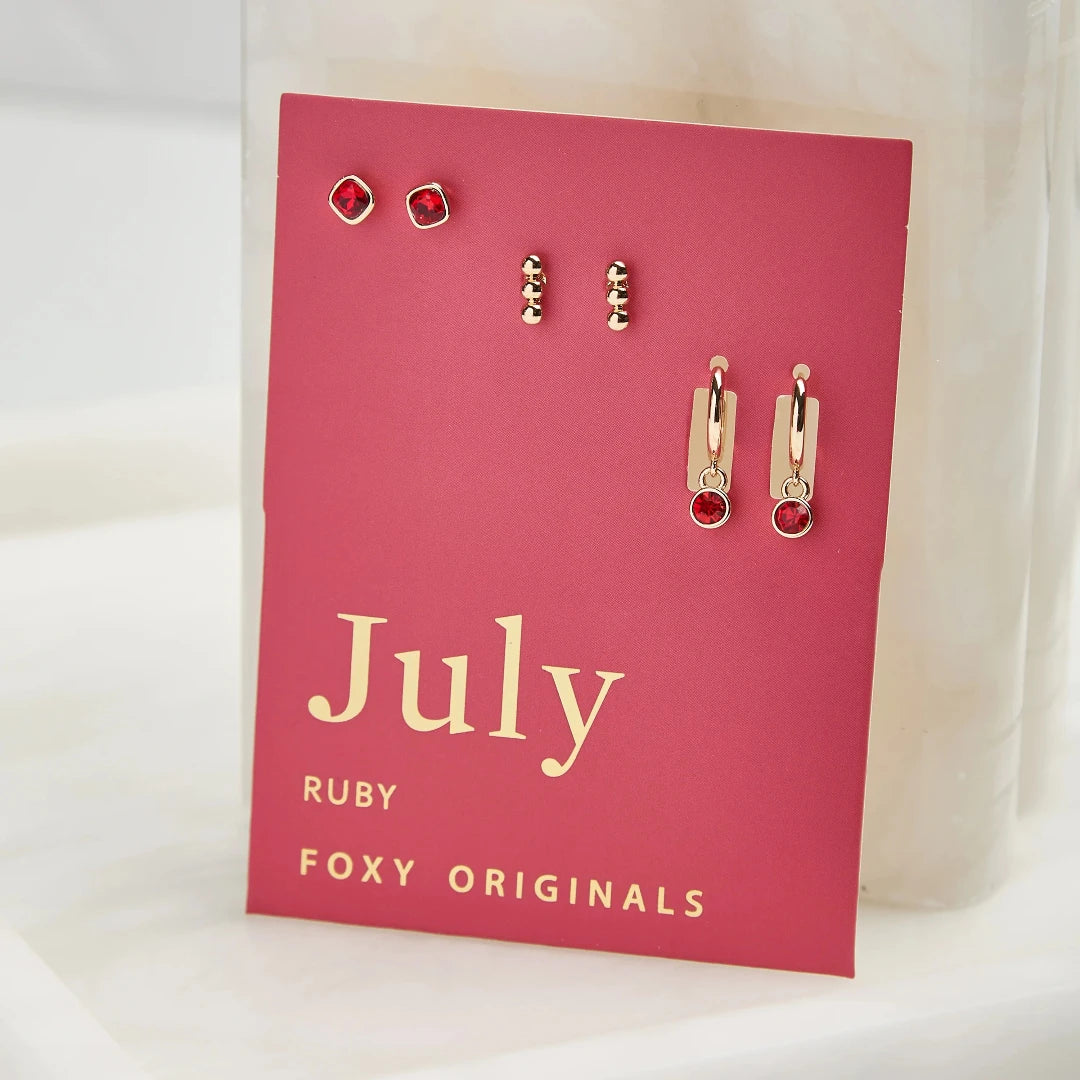 A set of three pairs of ruby earrings displayed on a red card. The set includes a pair of stud earrings, a pair of bar earrings with small ruby beads, and a pair of hoop earrings with dangling ruby stones. The card has the month "July" and the brand name "Foxy Originals."