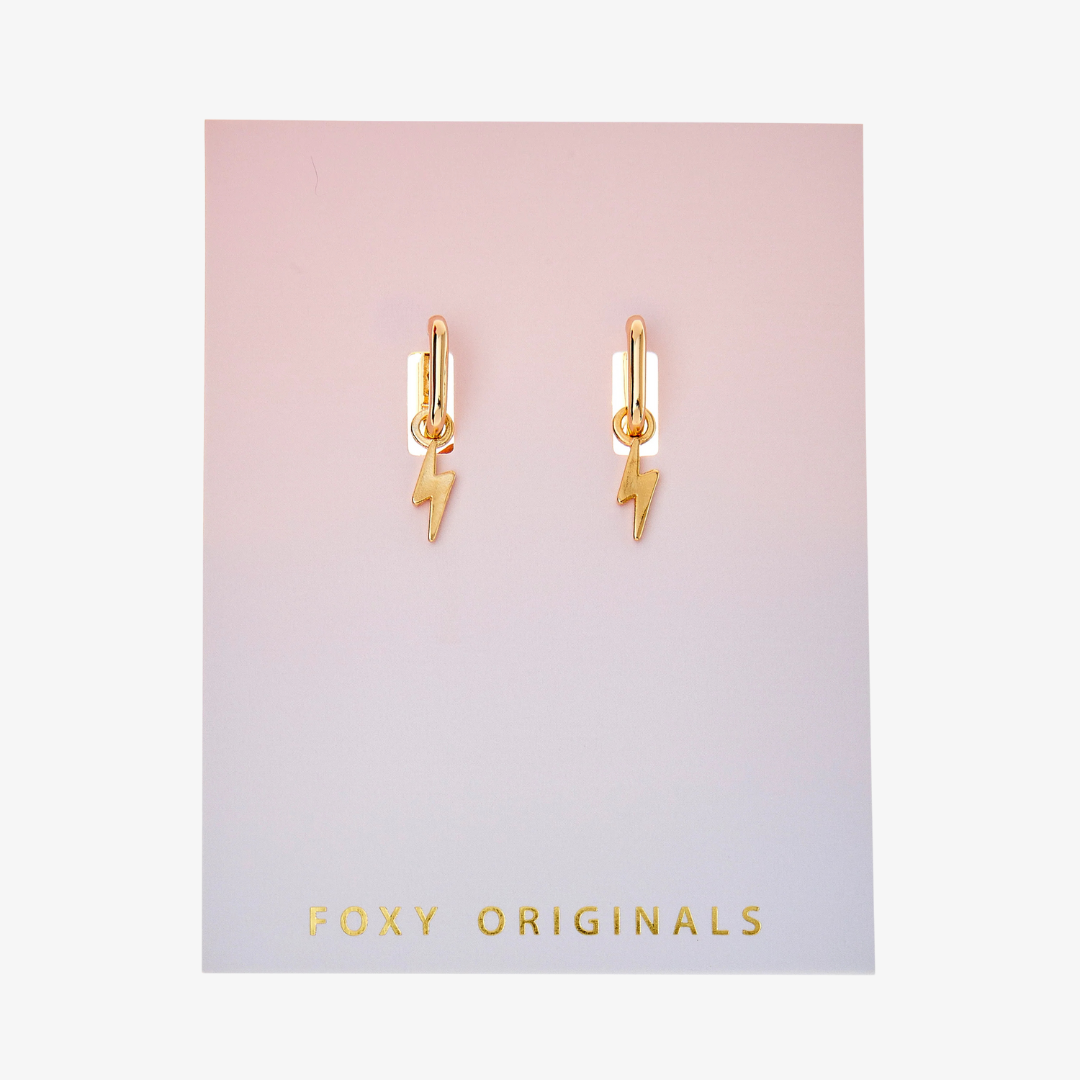 A pair of gold earrings with lightning bolt charms on a pink background. The earrings are displayed on a card with the brand name "Foxy Originals."