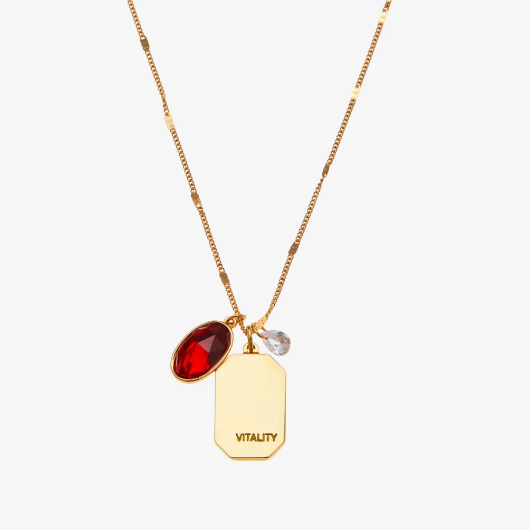 A gold necklace with a ruby stone pendant. The pendant also has the word "VITALITY" engraved on it. The necklace is displayed on a red card with the month "July" and the brand name "Foxy Originals."