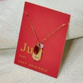 A gold necklace with a ruby stone pendant. The pendant also has the word "VITALITY" engraved on it. The necklace is displayed on a red card with the month "July" and the brand name "Foxy Originals."