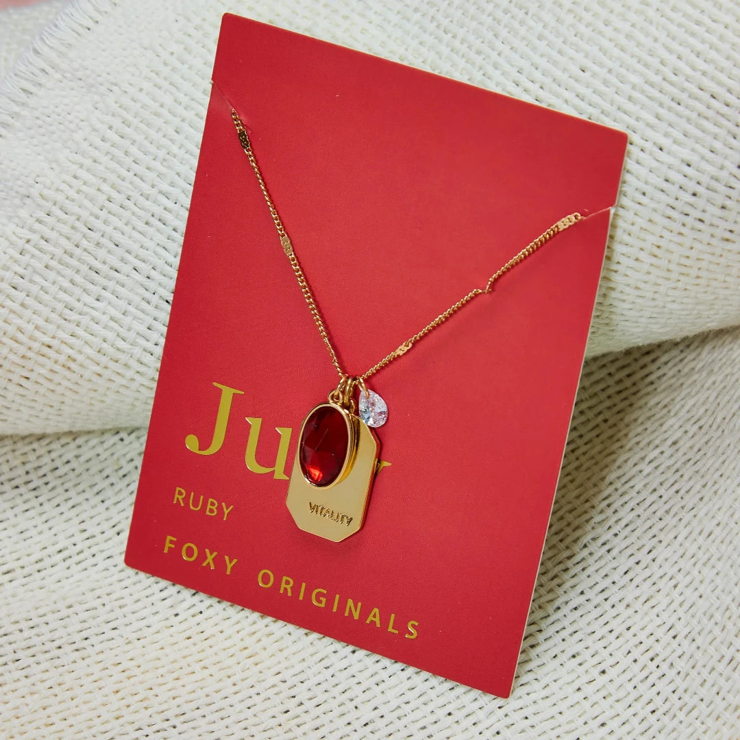 A gold necklace with a ruby stone pendant. The pendant also has the word "VITALITY" engraved on it. The necklace is displayed on a red card with the month "July" and the brand name "Foxy Originals."