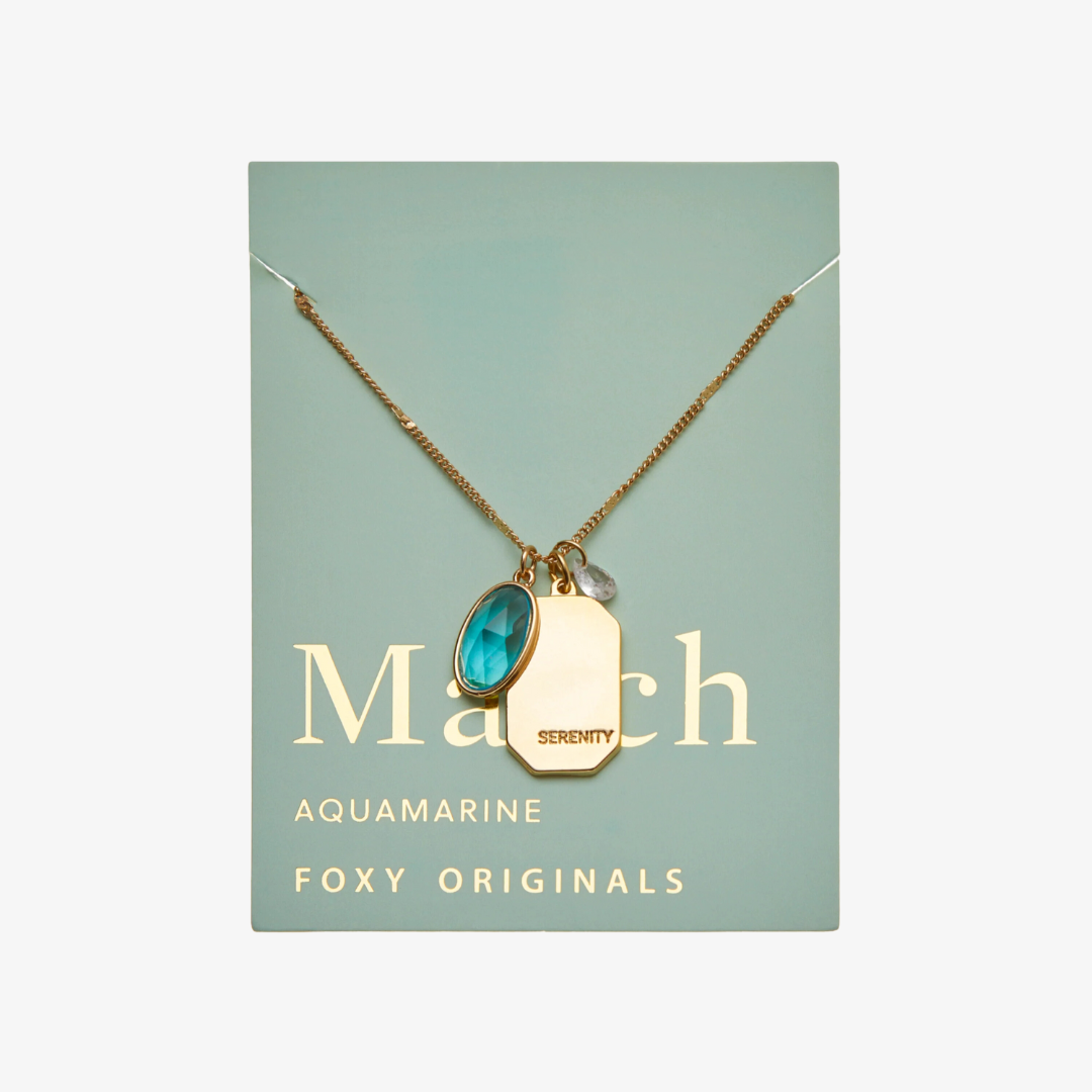 A gold necklace with an aquamarine stone pendant. The pendant also has the word "SERENITY" engraved on it. The necklace is displayed on a light blue card with the month "March" and the brand name "Foxy Originals."