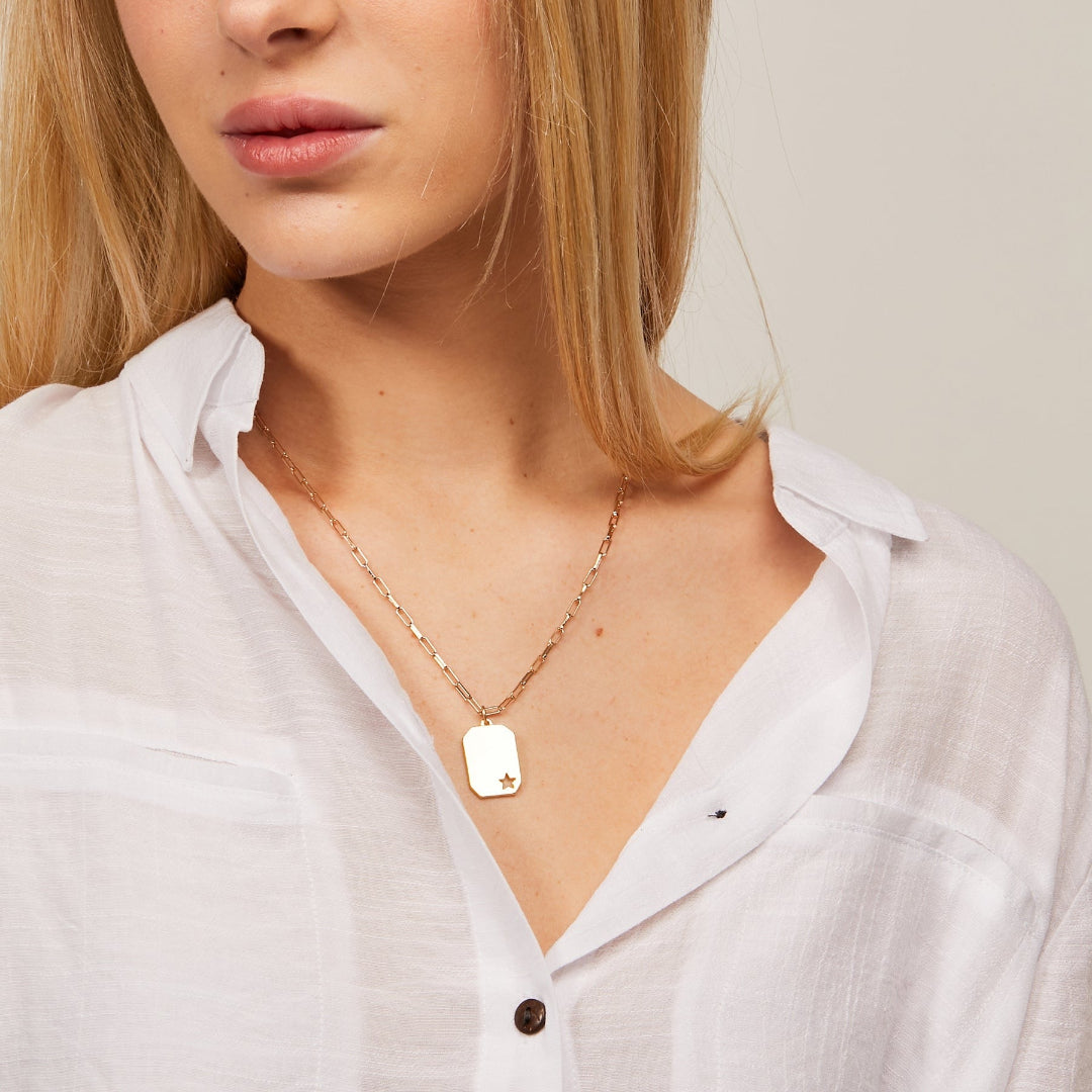 Person wearing a gold necklace with a tag pendant on a plain background