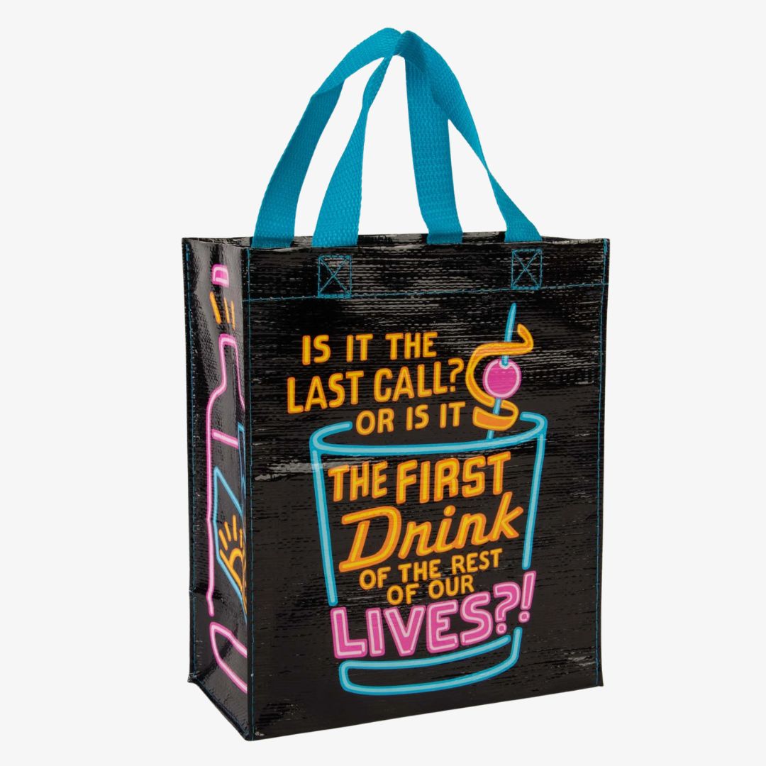 A black reusable tote bag with bright neon blue, pink, and yellow graphics. The front features a large cocktail glass with a cherry and straw, and text that reads "IS IT THE LAST CALL? OR IS IT THE FIRST DRINK OF THE REST OF OUR LIVES?!". On the left side, part of a neon bottle graphic is visible.