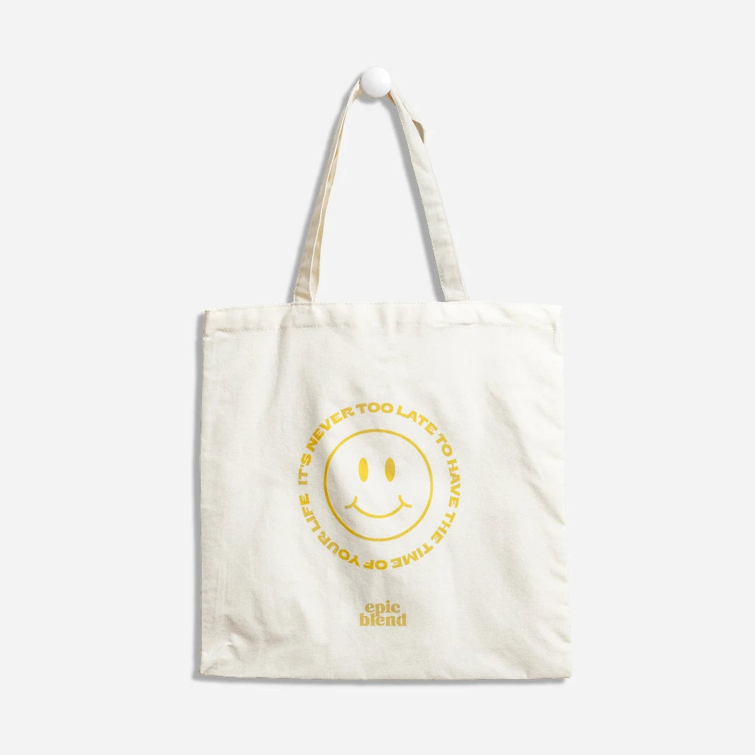 A white tote bag with a yellow smiley face design. The text around the smiley face reads: "It's never too late to have the time of your life." The bottom right corner displays the "Epic Blend" logo.