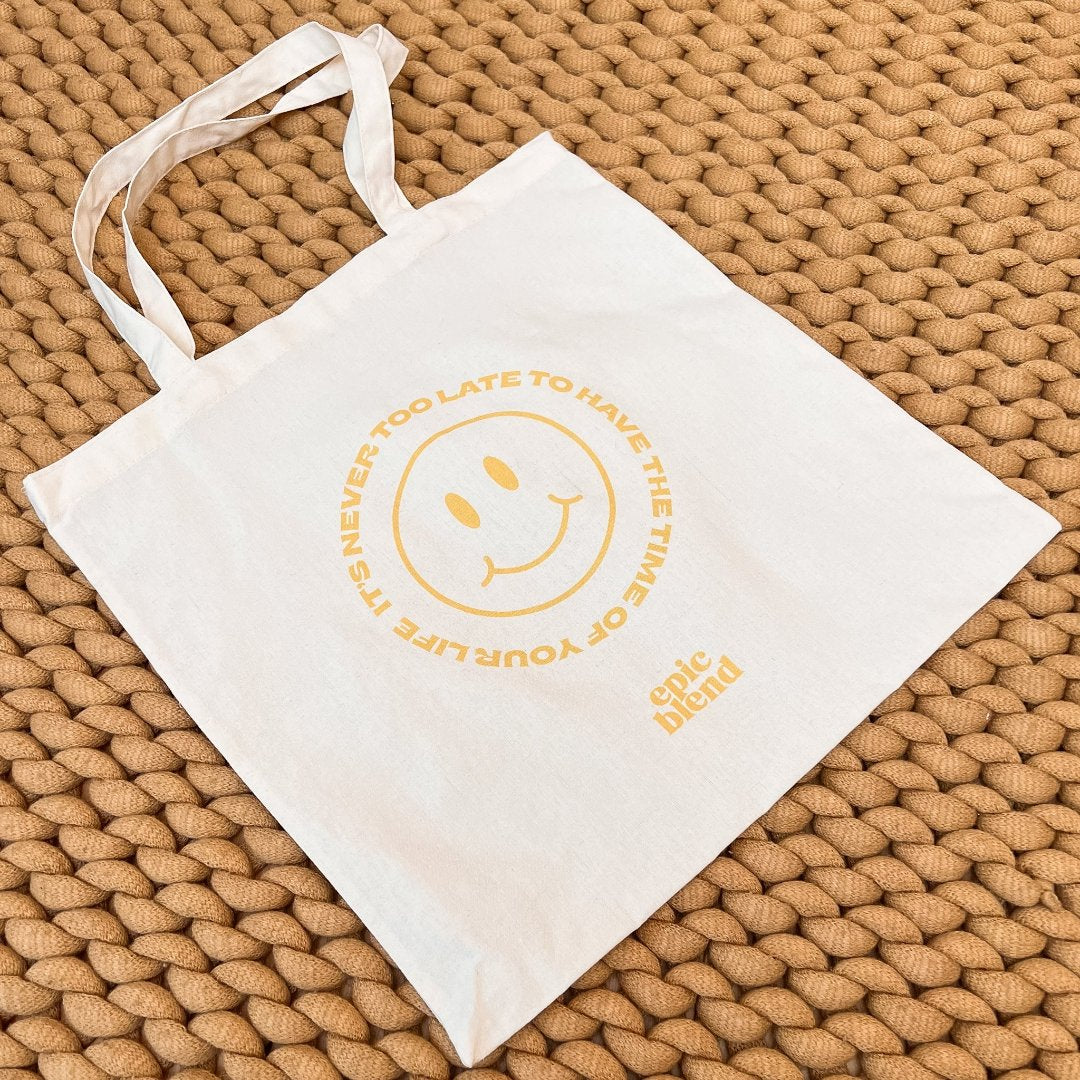 A white tote bag with a yellow smiley face design. The text around the smiley face reads: "It's never too late to have the time of your life." The bottom right corner displays the "Epic Blend" logo.