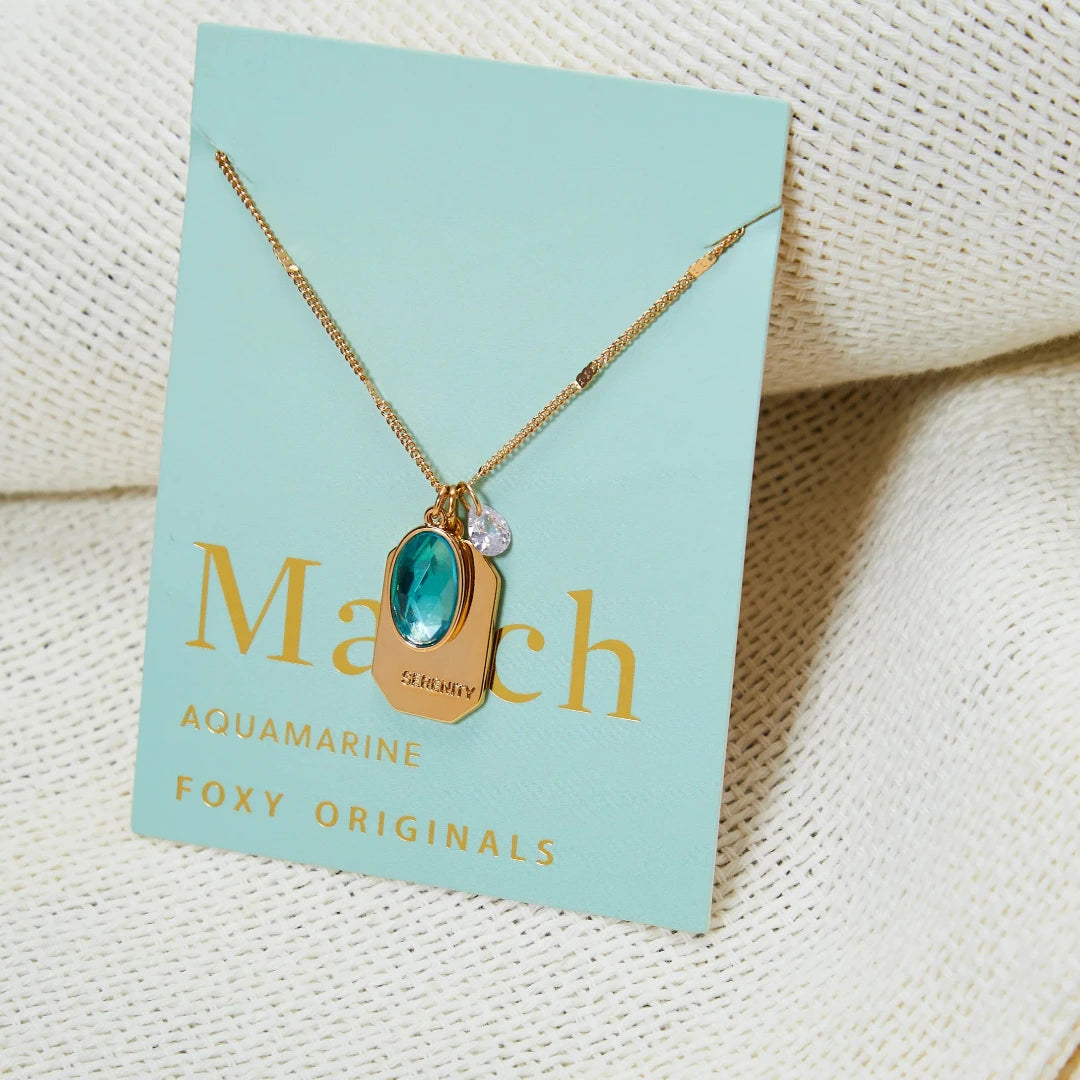 A gold necklace with an aquamarine stone pendant. The pendant also has the word "SERENITY" engraved on it. The necklace is displayed on a light blue card with the month "March" and the brand name "Foxy Originals."