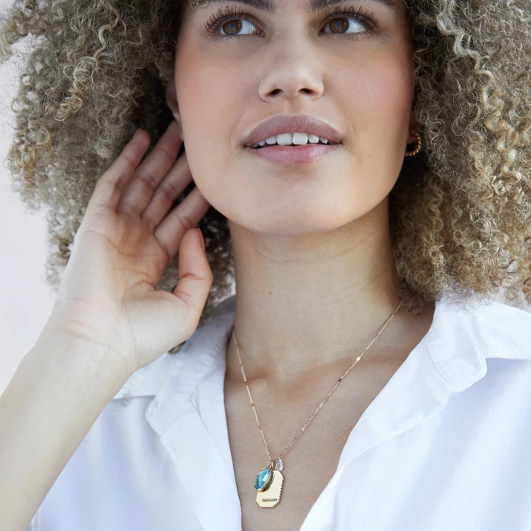 A gold necklace with an aquamarine stone pendant. The pendant also has the word "SERENITY" engraved on it. The necklace is displayed on a light blue card with the month "March" and the brand name "Foxy Originals."