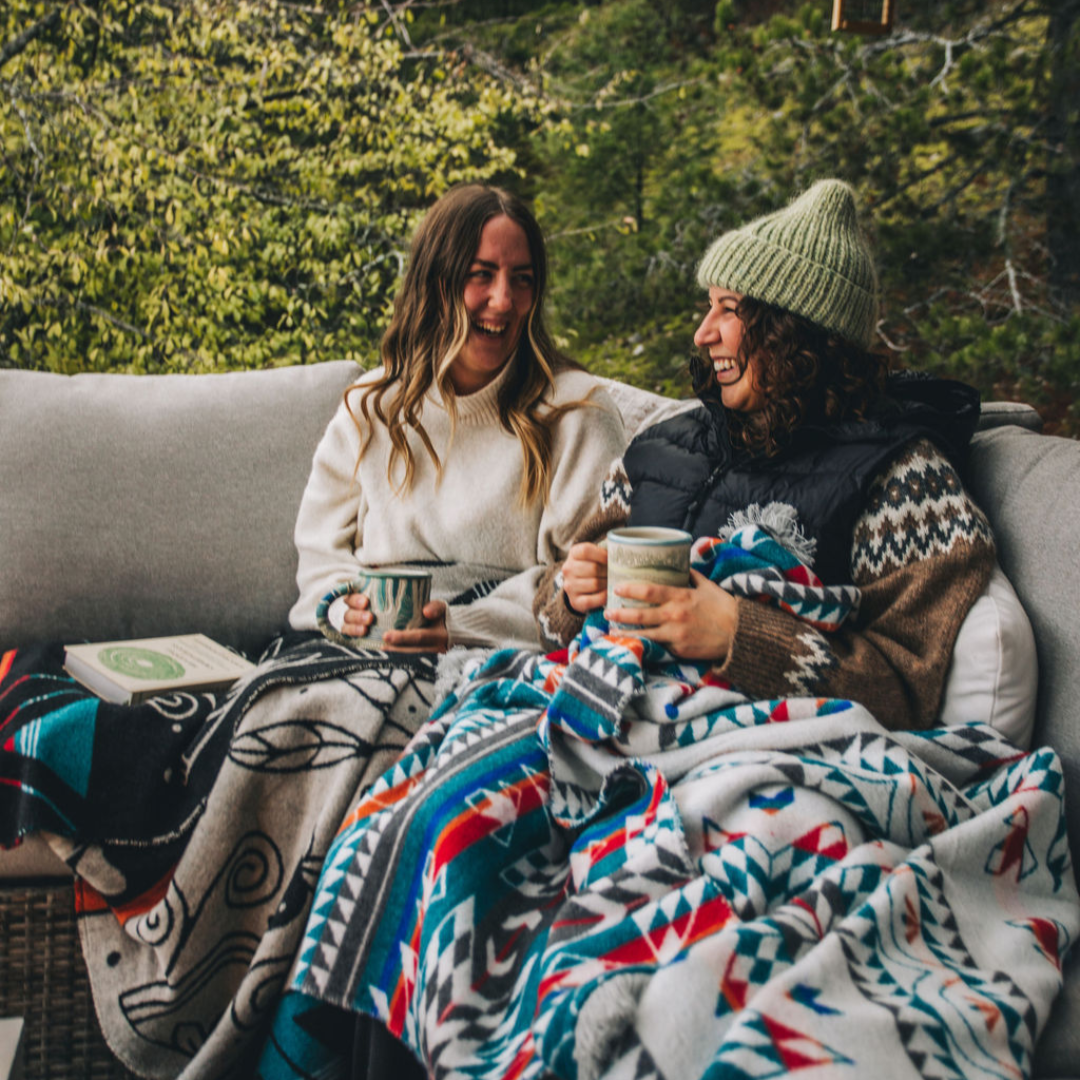 The Nibi Reversible Blanket by Mini Tipi, featuring bold Indigenous-inspired geometric patterns in teal, red, brown, and white, draped on the legs of two people sitting outside. This reversible and stylish blanket is available in Ottawa, Ontario, Canada.