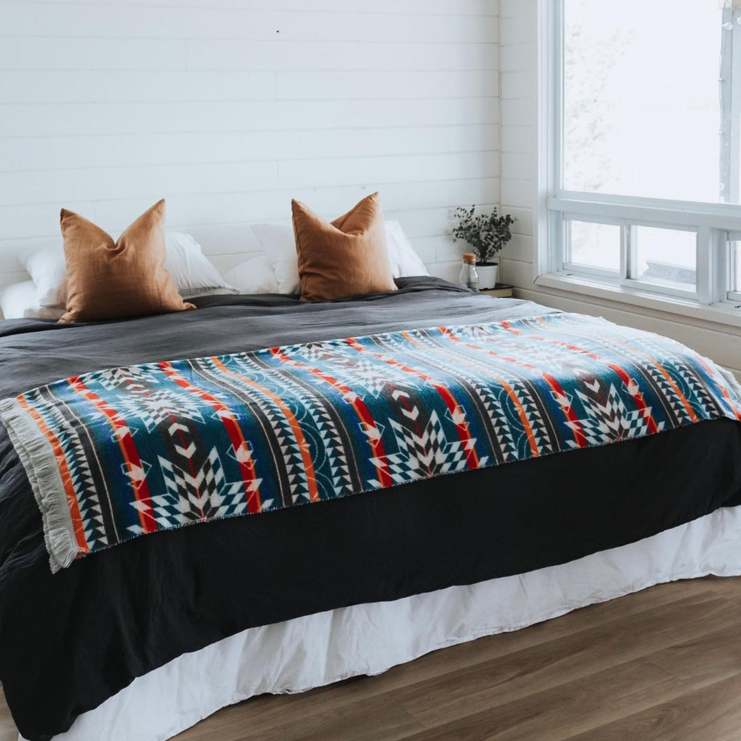 The Nibi Reversible Blanket by Mini Tipi, featuring bold Indigenous-inspired geometric patterns in teal, red, brown, and white, draped on a bed. This reversible and stylish blanket is available in Ottawa, Ontario, Canada.