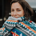 The Nibi Reversible Blanket by Mini Tipi, featuring bold Indigenous-inspired geometric patterns in teal, red, brown, and white, wrapped around one of the owners of MINITIPI. This reversible and stylish blanket is available in Ottawa, Ontario, Canada.