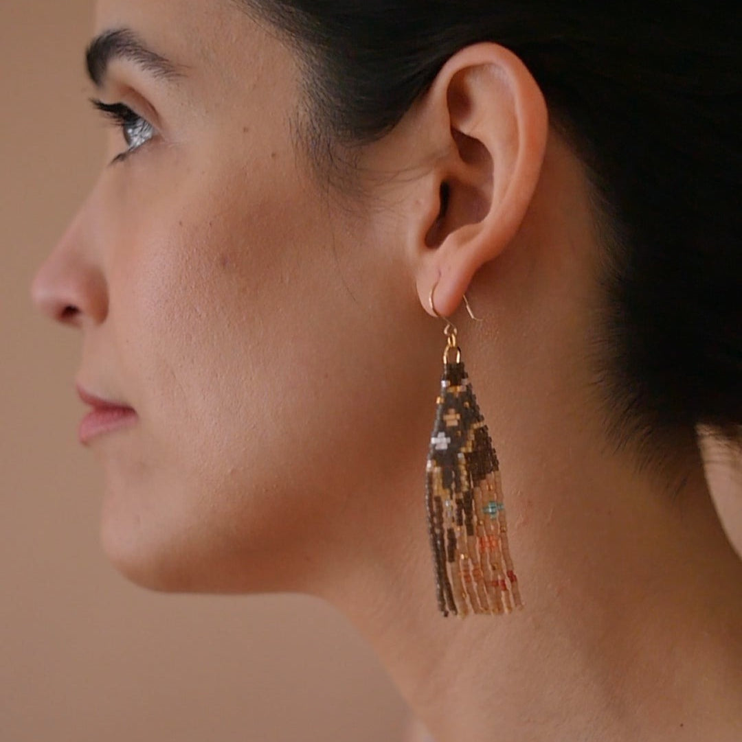 A pair of long, triangular tapestry-style earrings hand-stitched with small Japanese glass beads to depict a stylized face with light peach skin, blue eyes, coral cheeks, and a red mouth, framed by dark brown and grey hair with gold bead accents. They hang from delicate 14 KT gold filled fishhook ear wires.