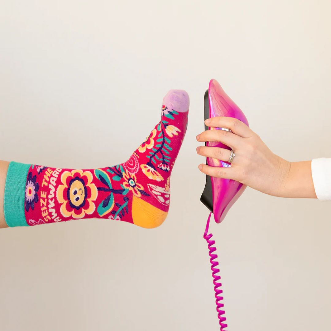 A pair of colourful women's crew socks with a red background featuring a whimsical floral pattern in shades of yellow, pink, green, and white. One sock displays the text "SEIZE THE AWKWARD" in white letters around a smiling yellow flower. The toe of the sock is light purple, the heel is yellow, and the cuff is bright green.