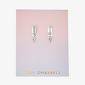 A pair of silver earrings with lightning bolt charms on a pink background. The earrings are displayed on a card with the brand name "Foxy Originals."