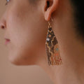 A pair of long, triangular tapestry-style earrings hand-stitched with small Japanese glass beads to depict a stylized face with light peach skin, blue eyes, coral cheeks, and a red mouth, framed by dark brown and grey hair with gold bead accents. They hang from delicate 14 KT gold filled fishhook ear wires.