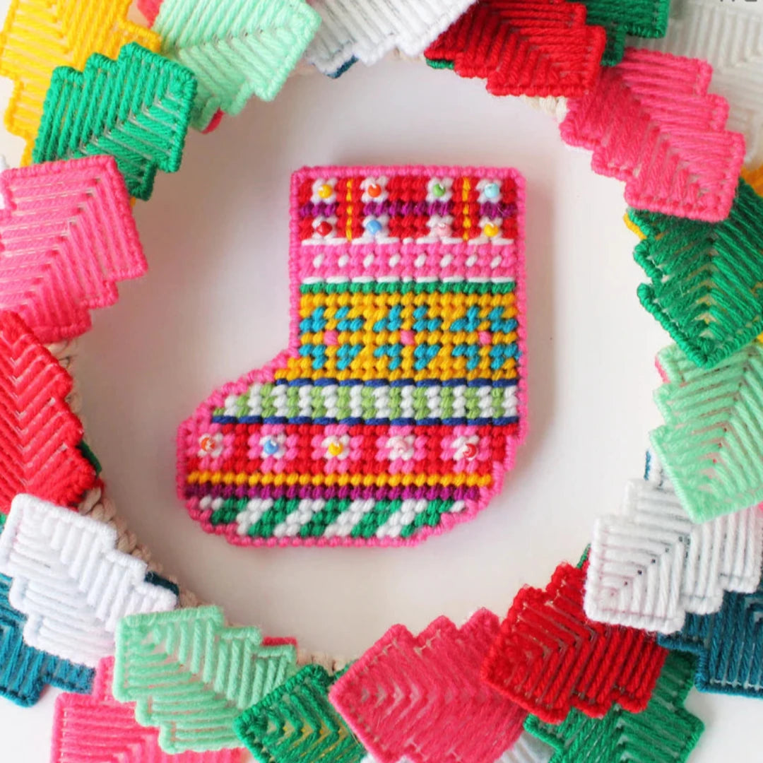 a colorful plastic canvas Christmas stocking ornament. The stocking is pink, yellow, blue, and green with white accents and colorful bead embellishments.