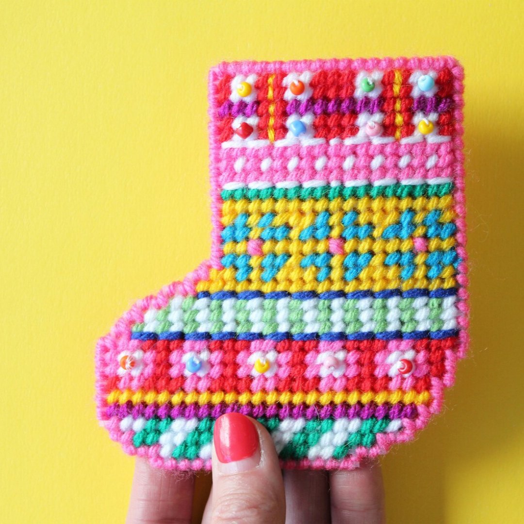 a colorful plastic canvas Christmas stocking ornament. The stocking is pink, yellow, blue, and green with white accents and colorful bead embellishments.