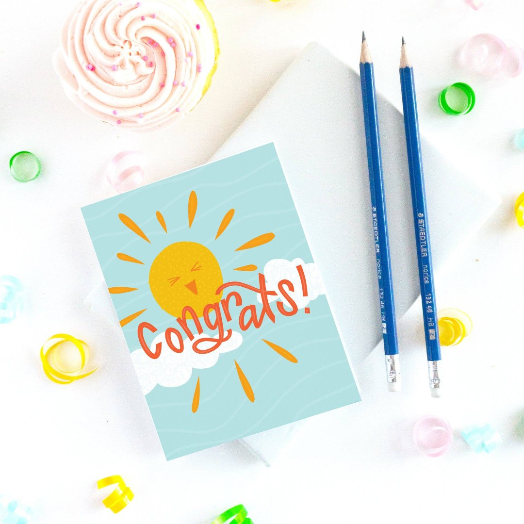 A congratulations card featuring a smiling sun with rays shining on clouds. The text "Congrats!" is written in bold letters below the sun. The card has a light blue background.