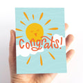 A congratulations card featuring a smiling sun with rays shining on clouds. The text "Congrats!" is written in bold letters below the sun. The card has a light blue background.