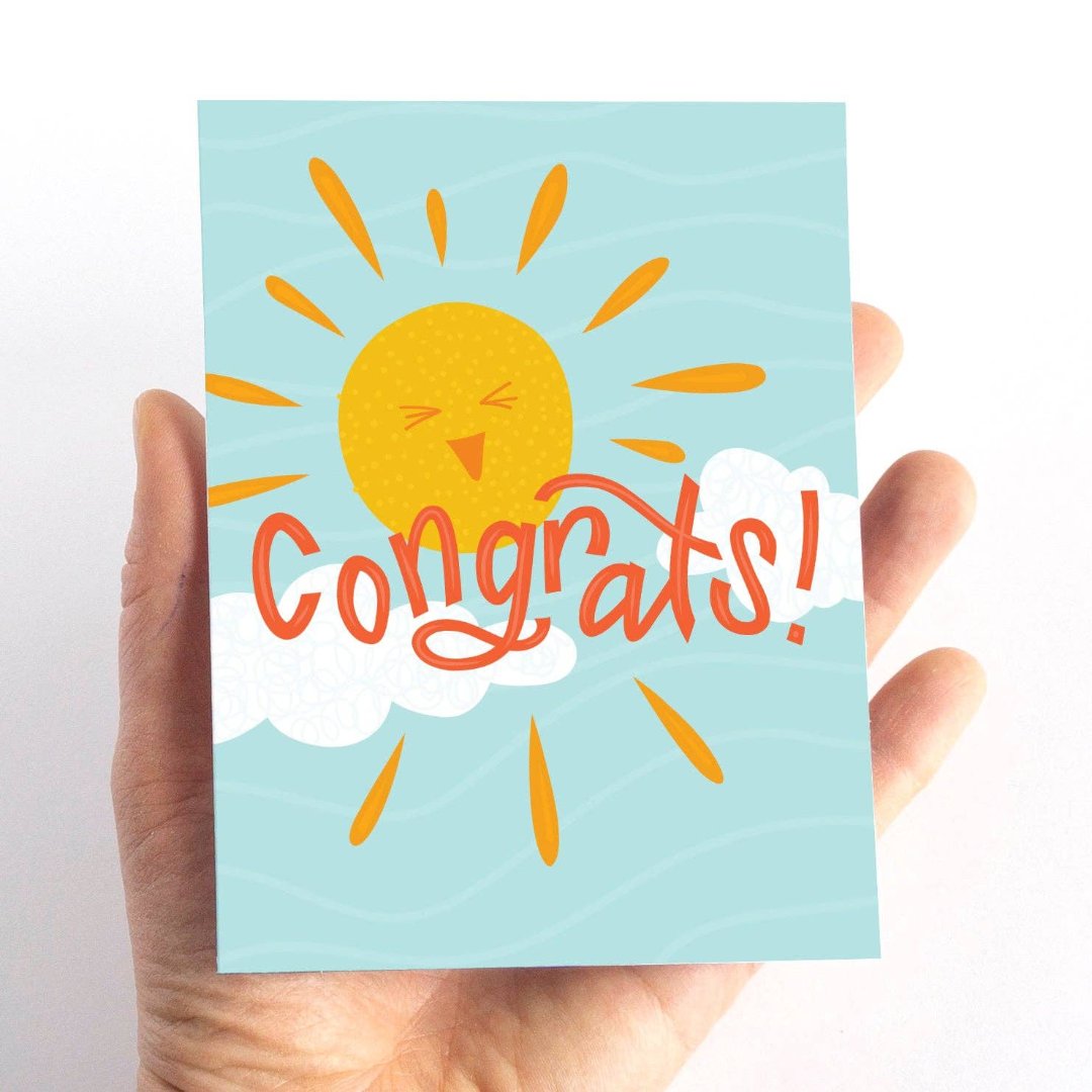 A congratulations card featuring a smiling sun with rays shining on clouds. The text "Congrats!" is written in bold letters below the sun. The card has a light blue background.