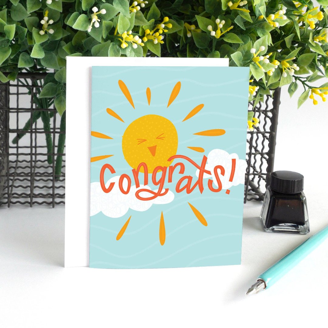 A congratulations card featuring a smiling sun with rays shining on clouds. The text "Congrats!" is written in bold letters below the sun. The card has a light blue background.
