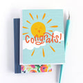 A congratulations card featuring a smiling sun with rays shining on clouds. The text "Congrats!" is written in bold letters below the sun. The card has a light blue background.