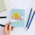 A congratulations card featuring a smiling sun with rays shining on clouds. The text "Congrats!" is written in bold letters below the sun. The card has a light blue background.