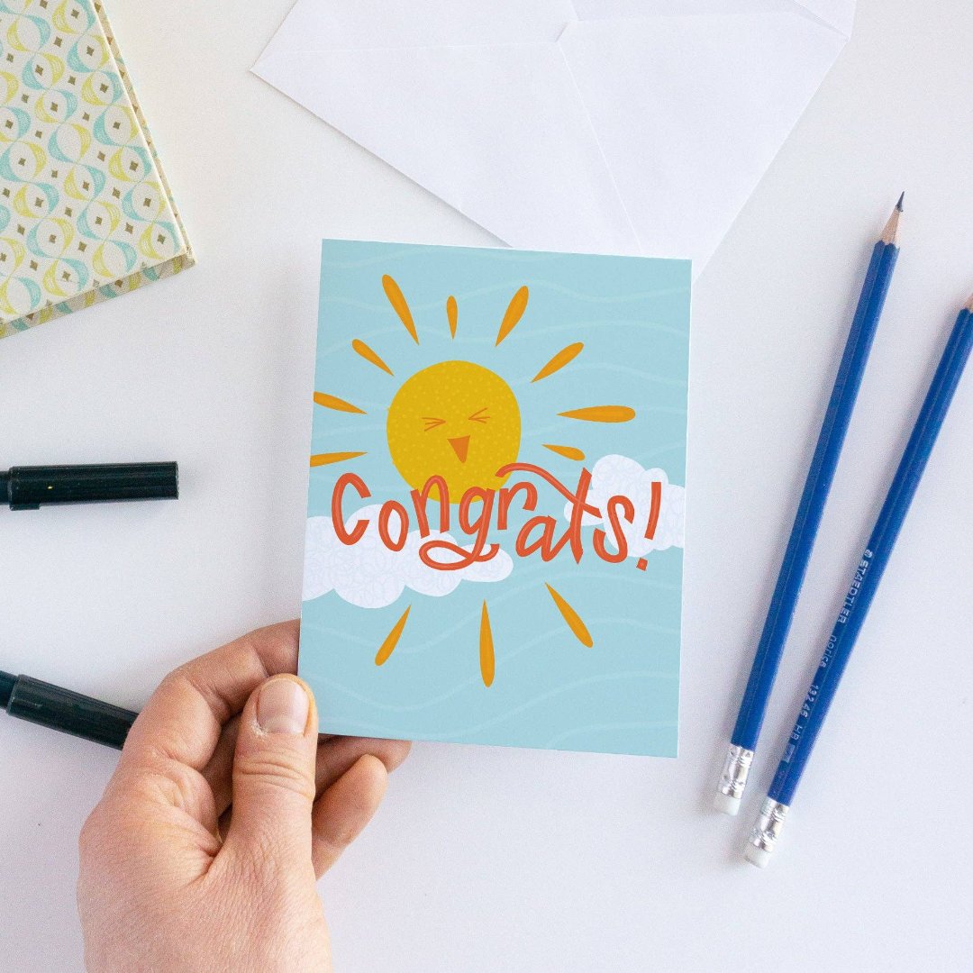 A congratulations card featuring a smiling sun with rays shining on clouds. The text "Congrats!" is written in bold letters below the sun. The card has a light blue background.