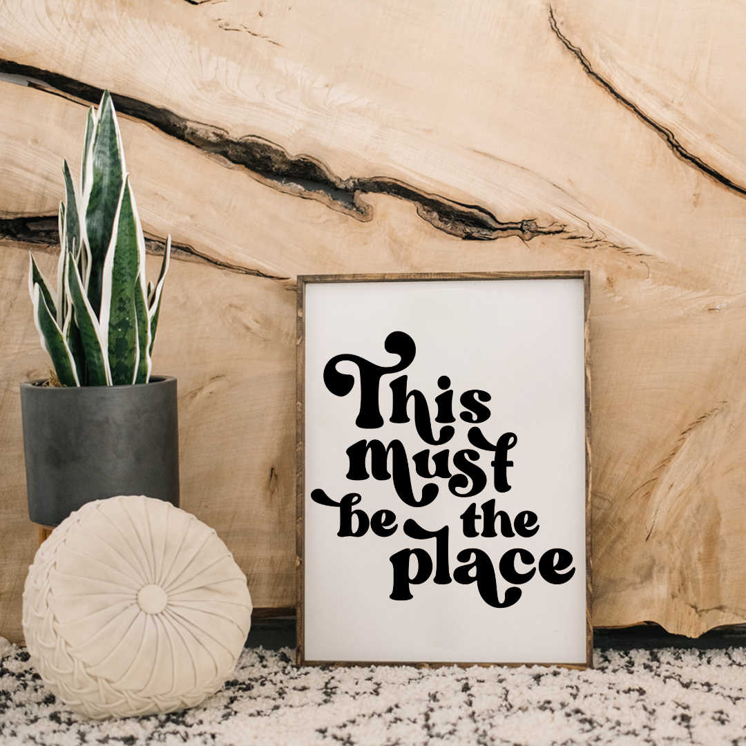 Decorative sign with 'This Must be the Place' text, potted plant, and round pillow against a wooden background.