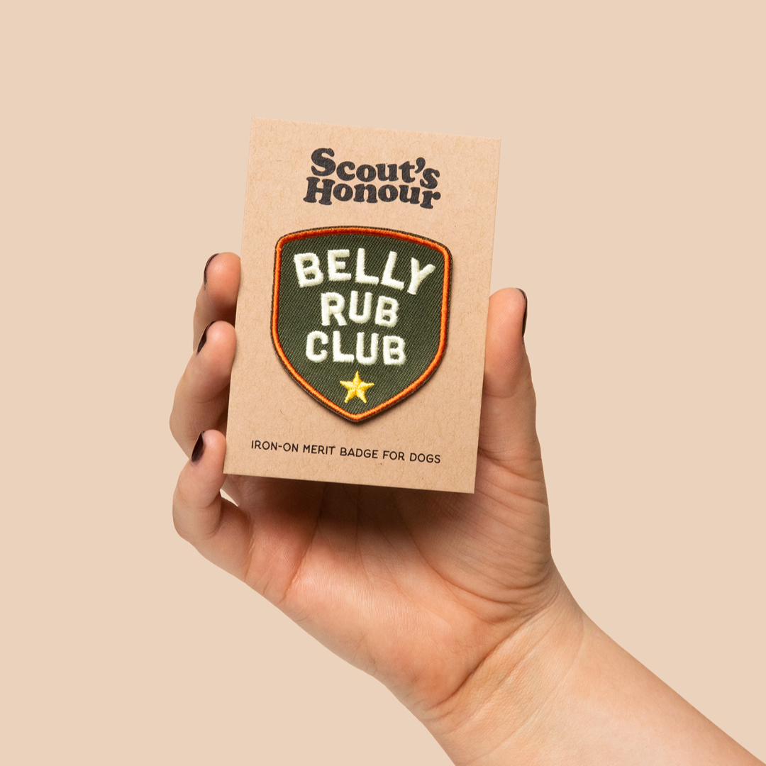Hand holding a 'Scout's Honour Belly Rub Club' product against a beige background