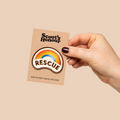 Hand holding a 'Scout's Honour' patch with a rainbow and 'Rescue' text on a beige background