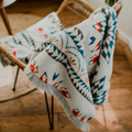 The Nibi Reversible Blanket by Mini Tipi, featuring bold Indigenous-inspired geometric patterns in teal, red, brown, and white, hanging from a chair. This reversible and stylish blanket is available in Ottawa, Ontario, Canada.