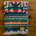 The Nibi Reversible Blanket by Mini Tipi, featuring bold Indigenous-inspired geometric patterns in teal, red, brown, and white, folded on a wood surface. This reversible and stylish blanket is available in Ottawa, Ontario, Canada.
